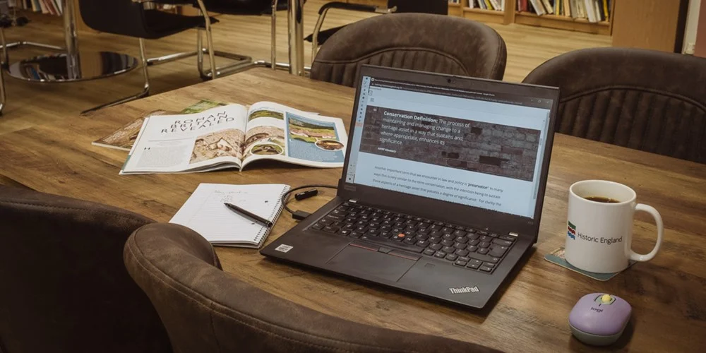 Develop skills for a career in heritage, at a pace that suits you A laptop on a desk with a magazine, a notepad and pen, a mug, and a mouse